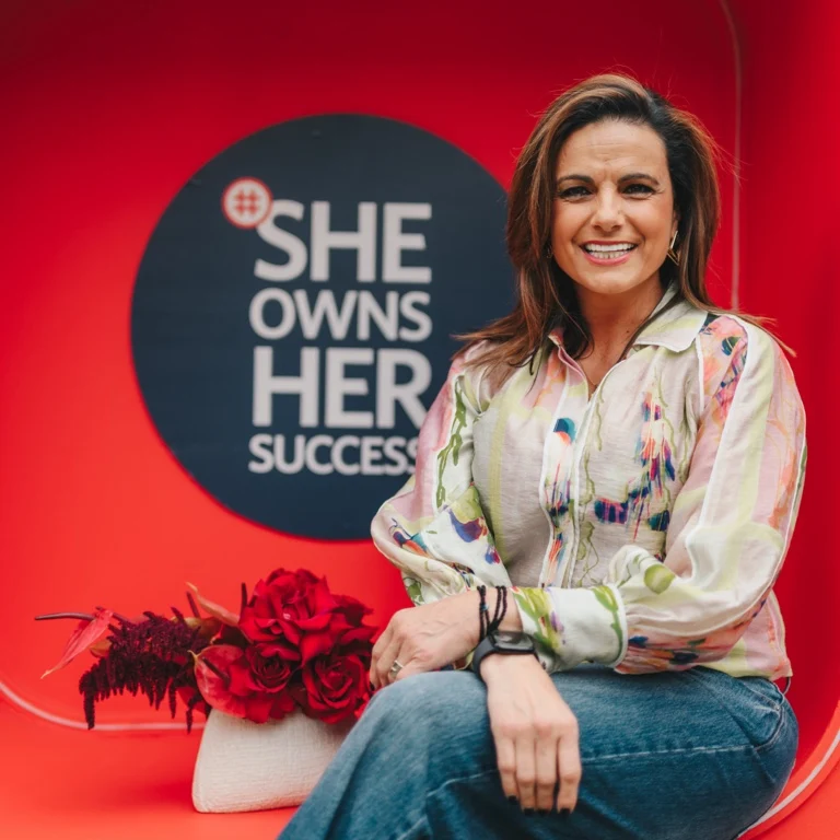 South African television presenter Leanne Manas sitting and smiling in front of a red background featuring a circular sign that reads 