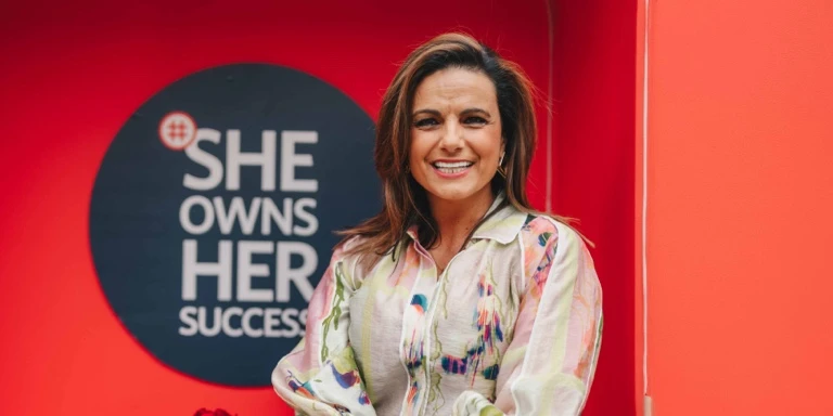 South African television presenter Leanne Manas sitting and smiling in front of a red background featuring a circular sign that reads 