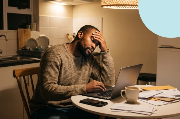 Stressed man sitting late at night at a kitchen table with a laptop and bills, illustrating the heavy burden of debt when not saving for medium to long term goals.