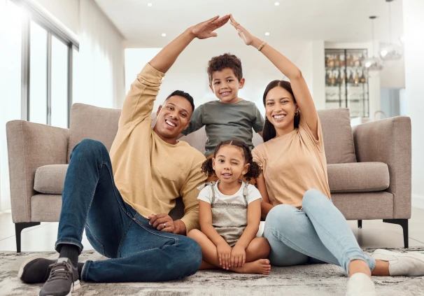 A smiling family of four sitting together in their living room, with the parents using their arms to form a protective roof shape over their children, representing the financial security and peace of 