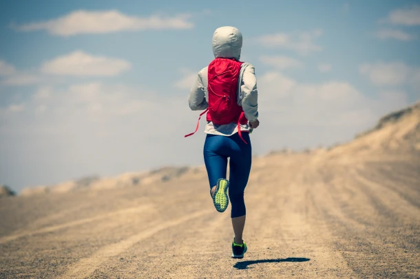 A woman jogging along a sandy road with a satchel on her back.