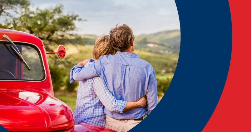 Senior couple embracing next to a red vintage car with scenic hills in the background, symbolising safe and enjoyable road journeys in South Africa.