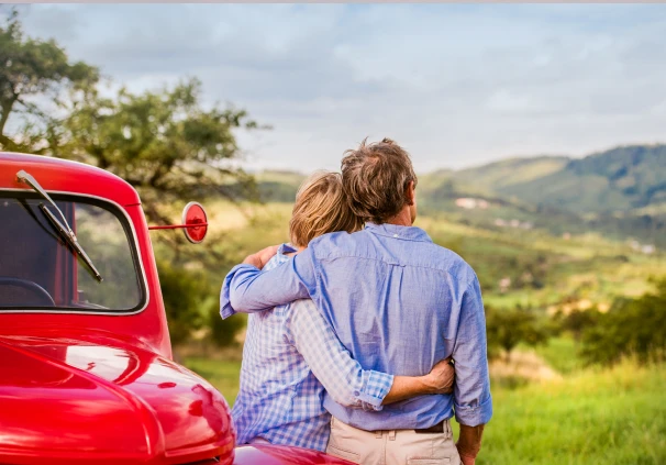 Senior couple embracing next to a red vintage car with scenic hills in the background, symbolising safe and enjoyable road journeys in South Africa.