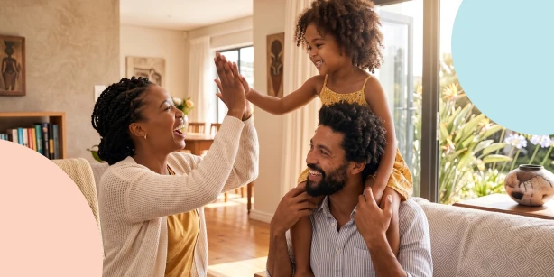A happy mother and father playing with their young daughter, who is sitting on her dad's shoulders and giving her mom a high five at home.