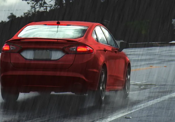 A red car on a wet road during heavy rainfall, with storm clouds overhead and trees in the background highlighting climate change and road safety awareness.