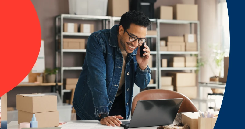 A freelance business owner smiling while managing his savings and tax obligations on a laptop and phone, surrounded by inventory and shipping boxes. 