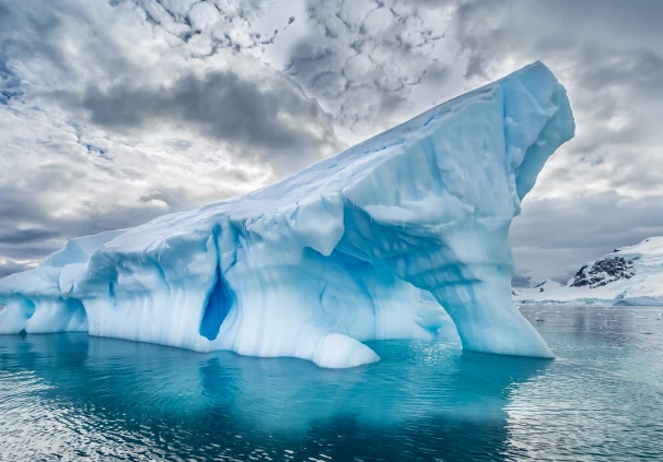 A dramatic polar iceberg with bright blue ice arching over clear turquoise waters, used as a visual metaphor to represent rising retirement costs.