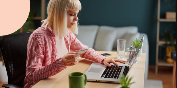 A Gen Z woman with blonde hair sitting at a wooden desk, holding a bank card while typing on her laptop, highlighting gen z’s online financial habits.