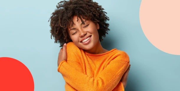 A joyful young woman with curly hair hugging herself against a blue background, reflecting the peace of mind from goal-setting habits that achieve financial security.