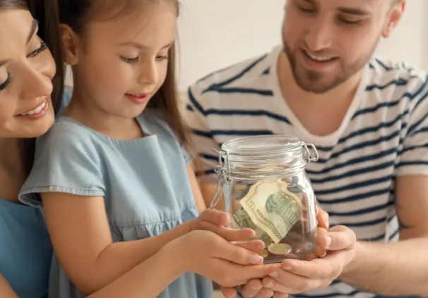 Parents on either side of their young daughter, looking inside a small savings kitty, teaching her the importance of early saving. 