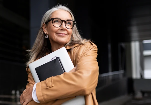 A smiling, confident and successful senior businesswoman holding a laptop, conveying the wisdom of timeless money lessons.