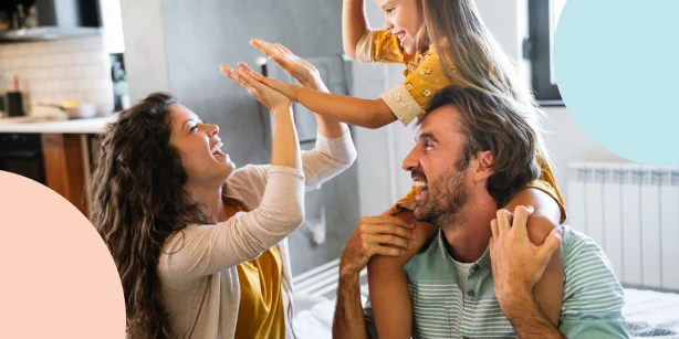 A happy mother and father playing with their young daughter, who is sitting on her dad's shoulders and giving her mom a high five at home.