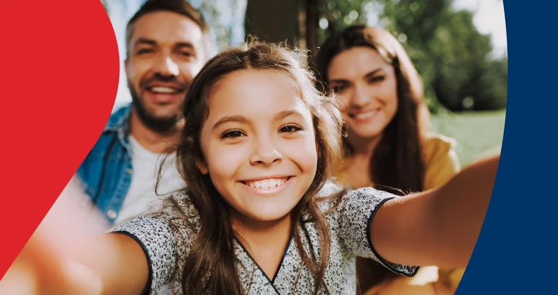 A smiling family of four sitting together in their living room, with the parents using their arms to form a protective roof shape over their children, representing the financial security and peace of mind that estate planning provides.