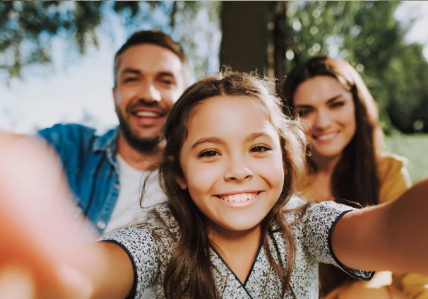 Smiling young girl taking a selfie outdoors with her parents in the background. Her parents have secured their legacy with an executable will to ensure she is supported if something happens to her.