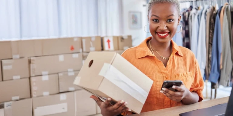 Smiling female business owner in an orange shirt holding a package and using a smartphone in a boutique clothing store. Shelves with boxes and racks of clothes suggest a thriving retail business, secured with business insurance.