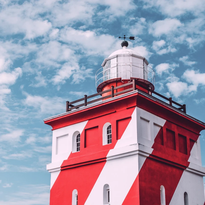  Red-and-white striped lighthouse with a glass lantern room on top, set against a bright blue sky with scattered clouds.
