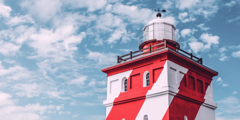  Red-and-white striped lighthouse with a glass lantern room on top, set against a bright blue sky with scattered clouds.