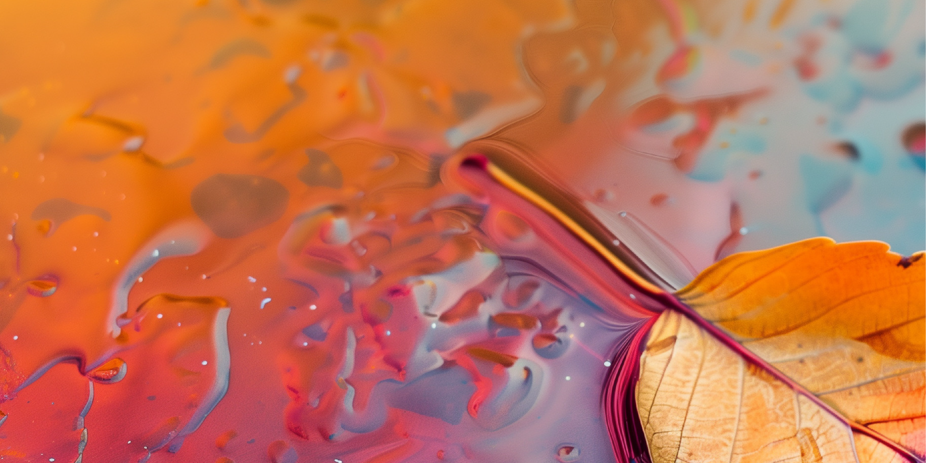 A close-up overhead view of a yellow leaf resting in a shallow puddle of water with swirling, iridescent reflections of orange, pink, and blue. 