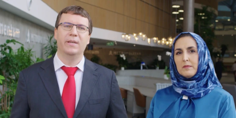  Two people standing indoors in a modern lobby or office atrium, facing the camera. One person wears a dark suit with a red tie and glasses, while the other wears a blue headscarf and outfit.
