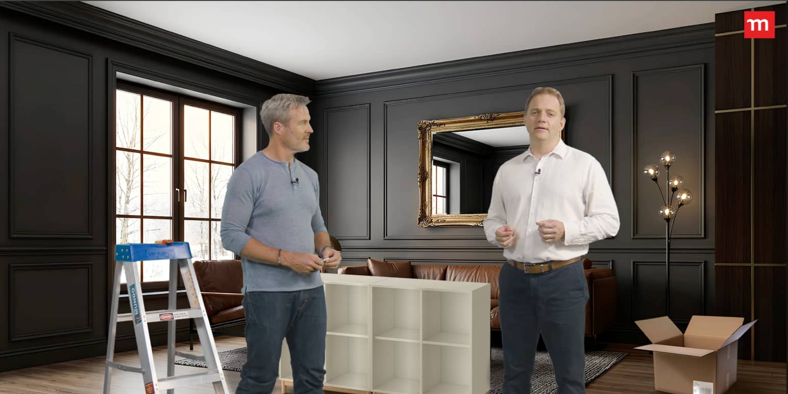 Two men standing in a stylish dark-paneled living room, discussing furniture assembly beside a ladder, shelving unit, and open box.