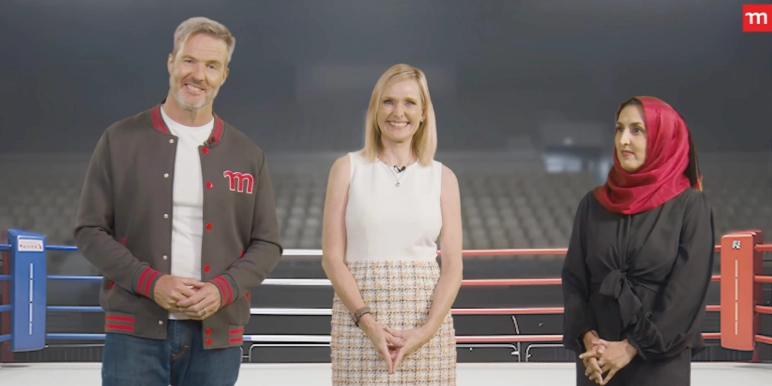 Three individuals stand in front of a boxing ring setup inside a large, empty stadium or arena.