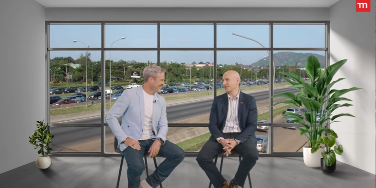 Two men in business attire sit on stools in a modern studio with floor-to-ceiling windows overlooking a busy highway and parking lot, engaged in conversation, with large potted plants on either side.