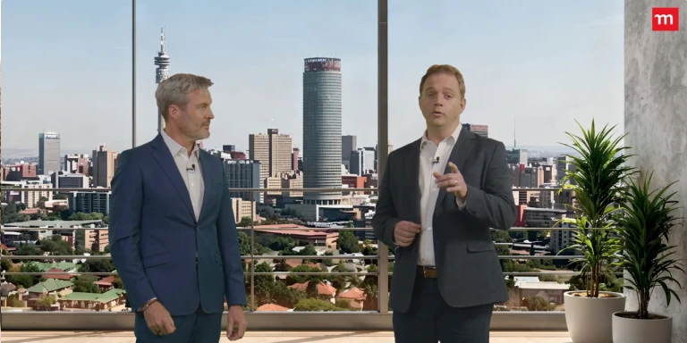 Two men in business suits stand in a modern studio with a large window overlooking a city skyline, as one speaks and gestures toward the camera while the other looks on.