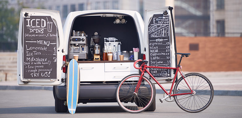 Back of a panel van that's converted into a mobile coffee shop with a surf board and a bicycle placed next to it. 