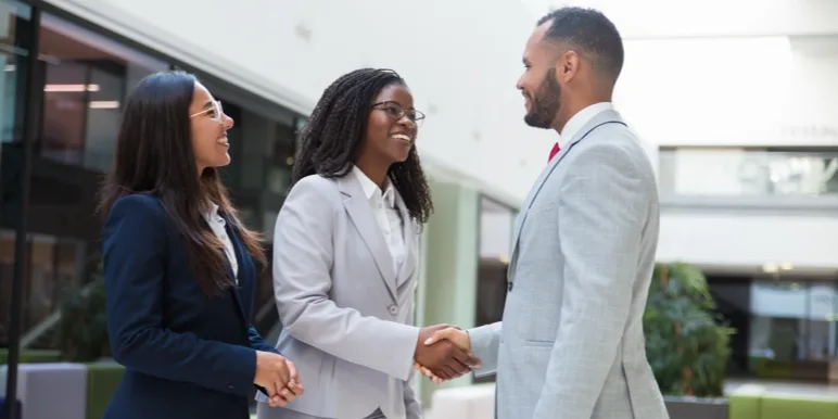 Two women smiling at each other and shaking hands inside an office building.