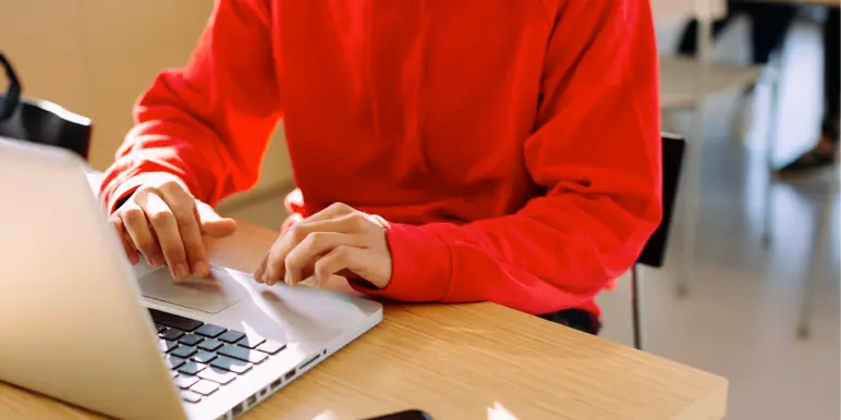 A person in a red hoodie using a laptop at a wooden desk to process two-pot online withdrawals.