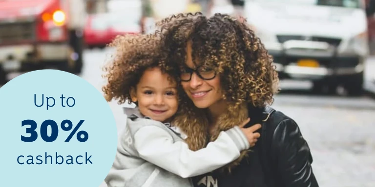 A woman and her young daughter smiling and kneeling down in front of a busy roadside background with cars. Get up tp 30% cash back on your yearly premiums.