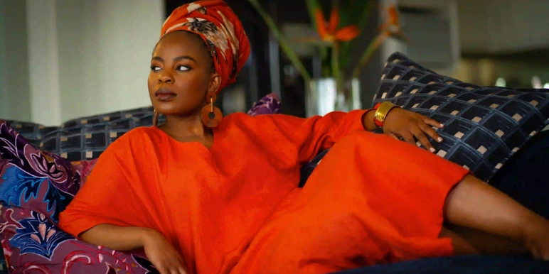 A lady lounging on a couch wearing a bright orange casual dress and patterned headwrap, with colorful cushions and flowers in the background.