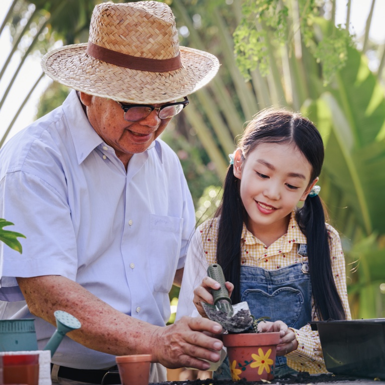 A grandfather wearing a straw hat helping his granddaughter with some gardening. 