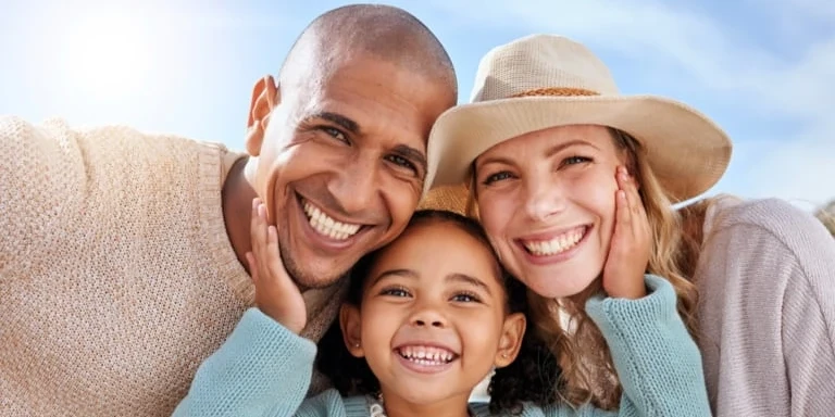 Parents enjoy a day outdoors with their young daughter. The daughter is standing in the middle of them with her hands on each parent's cheeks.