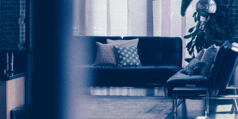 Modern living room with dark sofas, patterned throw pillows, a large rug, and indoor plants near tall windows.