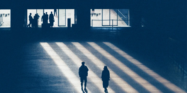 Two people walking across a large open indoor space with long shadows cast by sunlight streaming through windows, while a group stands in the background near the entrance.