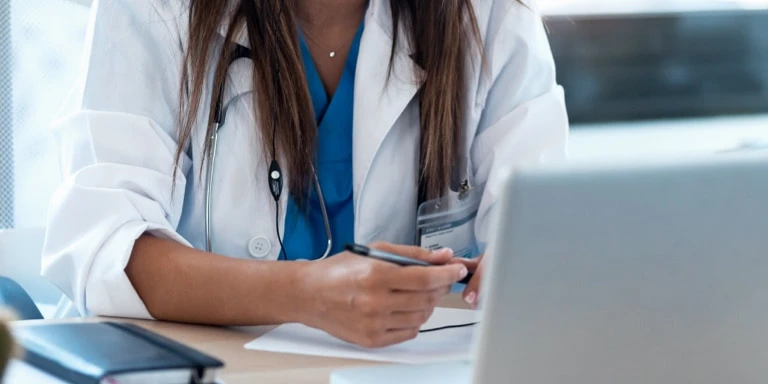 Close-up of a female medical professional wearing a stethoscope and white coat, writing notes at a desk in front of an open laptop.