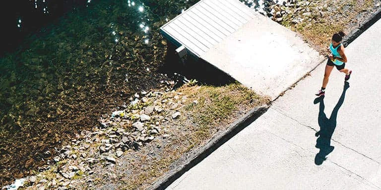 Aerial view of a person jogging along a paved path beside clear water and a small wooden dock on a sunny day.