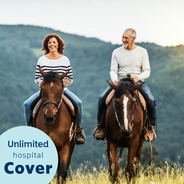 An elderly woman and man, happily riding horses outdoors with mountains in the background.