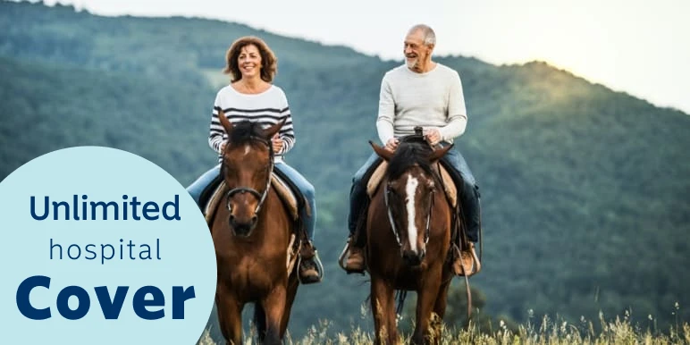 An elderly woman and man, happily riding horses outdoors with mountains in the background.