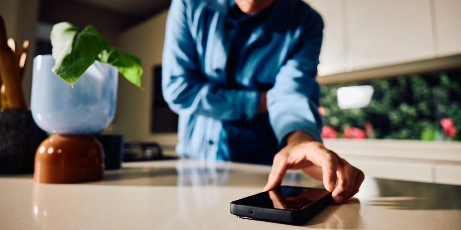 A person standing in a kitchen reaching toward a smartphone requiring emergency assistance at home.]