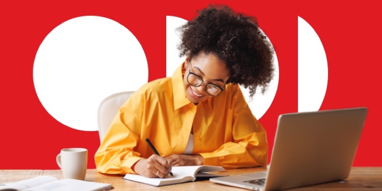 A woman seated at a desk as she writes down her savings goals in a notebook with her laptop in front.