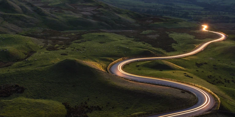 An aerial view of a winding road illuminated by evening light trails, illustrating the patience and planning required to build the future you want.
