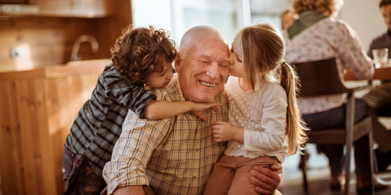 Elderly person smiles while his two grandchildren kiss his cheeks on either side. Indoors, warm setting. One child has long hair, the other curly. A third person is partially visible in the background.