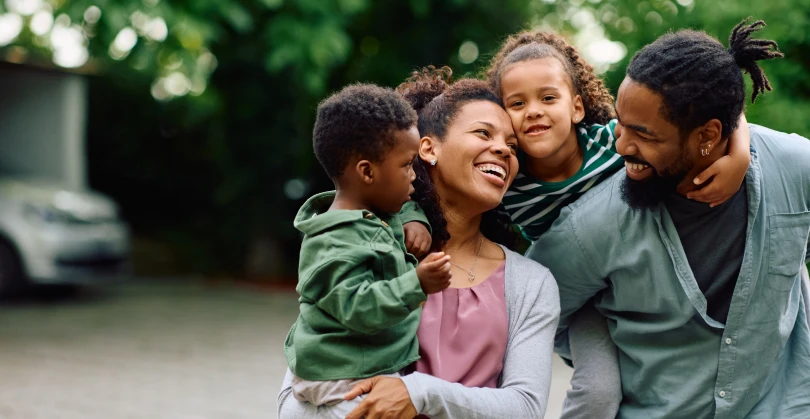 A happy Black family standing outside near a driveway; a mother smiles while holding a young boy in a green jacket, while a father with dreadlocks laughs as he carries a young girl piggyback style on his back