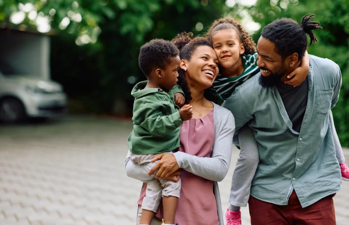 A happy Black family standing outside near a driveway; a mother smiles while holding a young boy in a green jacket, while a father with dreadlocks laughs as he carries a young girl piggyback style on 